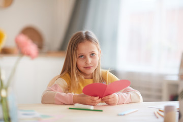 Portrait of cute little girl holding heart-shaped card and looking at camera while sitting at table in cozy home interior, copy space