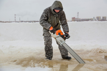 Worker in a green jacket with a hood with a chainsaw in his hands