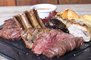 Plate of meat, chicken, ribs, black pudding, sausage, potatoes and sauce on black stone plate