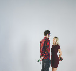 Charming couple love in red tone outfits standing, smiling ​and looking each other. A man wears eyeglasses​ hides red rose and a woman hides gift box​ behind own back, on white background. 