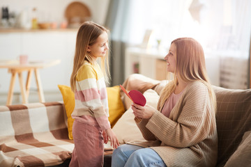 Side view portrait of cute little girl giving handmade card to mom on Mothers day in cozy home interior