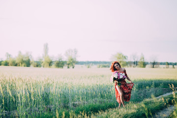 Naklejka na ściany i meble Red-haired woman in a red dress with a bouquet of peonies walking along a path in a summer field at sunset.