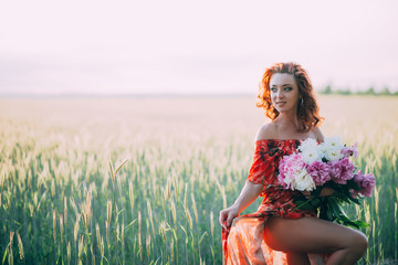 Red-haired woman in a red dress with a bouquet of peonies flowers dancing in a wheat field in summer at sunset. Soft focus. Back light.