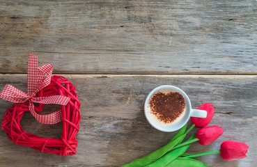 Romantic coffee break concept. Cup of coffee with heart foam, bouquet of red tulips and wicker red heart on old wooden background. Space for text, top view