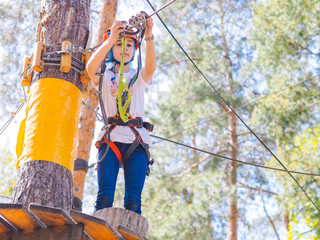 Teenager in orange helmet climbing in trees on forest adventure park. Girl walk on rope cables and high suspension bridge in adventure summer city park. Extreme sport equipment helmet and carabiner.