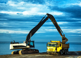 backhoe or digger working with bucket at industrial earth excavation site loading dump truck in sunrise light