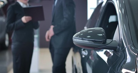 Unrecognizable Caucasian trader and client signing sale and purchase agreement in auto showroom. Focused on black car standing at the foreground. Selling, purchasing, industry. Cinema 4k ProRes HQ.