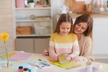 Warm-toned portrait of happy mother hugging daughter while drawing pictures at wooden kitchen table, copy space