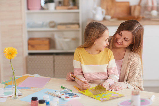Warm-toned portrait of happy mother hugging daughter while painting pictures at wooden kitchen table, copy space