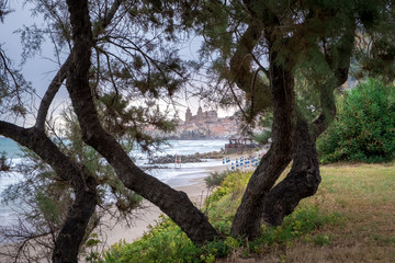 Fototapeta premium Seafront of Tyrrhenian Sea. View of the Sicilian city of Cefalu through trees growing on the shore