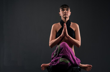 Obraz premium Front view of charming girl sitting in lotus position with prayer hands. Pretty lady looking up and meditating. Isolated on black background with copy space. Concept of yoga practice.