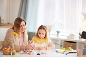 Warm-toned portrait of smiling mature mother painting pictures with cute little daughter while sitting at wooden table in cozy home interior, copy space