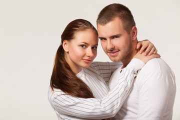 Front view of charming young woman hugging joyful guy from behind and biting ear. Handsome man holding girlfriend hand and smiling. Isolated on white studio background. Concept of relationships.