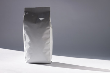 Blank white foil bag on monochrome background in minimal style with natural light shadow. Packaging for foods or drinks with valve and seal template mockup. Metallic coffee tea retail package design.