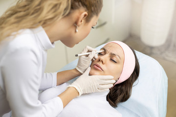Fototapeta premium Marking eyebrows with a pencil. Permanent makeup, tattooing of eyebrows. Cosmetologist in white gloves applying make up for woman in beauty salon