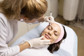 Marking eyebrows with a pencil. Permanent makeup, tattooing of eyebrows. Cosmetologist in white gloves applying make up for woman in beauty salon