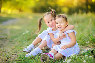 Fototapeta premium Cute children are walking in the park at sunset. Girls hug while sitting on the grass and laugh cheerfully. Children play and have fun outdoors.