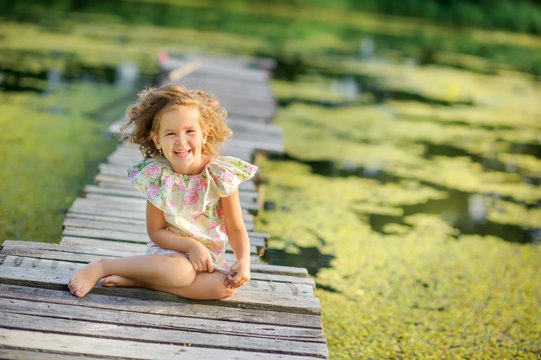 Happy girl sits and rests on a wooden pier on the lake. Children have a rest outdoors. Childhood.