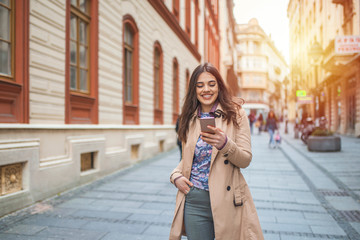 Obraz premium beautiful woman is sending a text message using an app on her smartphone while walking in the street on a sunset background. Happy brunette woman typing sms on smartphone in city