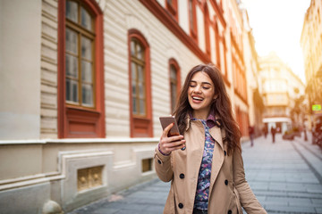 Gorgeous beautiful young woman with brown hair messaging on the smart-phone at the city street background. pretty girl having smart phone conversation in sun flare.