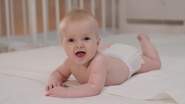 Smiling little baby girl with big blue eyes lying on her tummy on white bed looking at camera and smiling. happy baby in white lies on the bed and touches the legs with his hands.