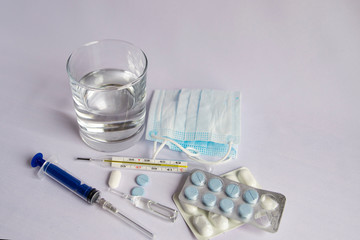 A glass of water, a thermometer with a high temperature indicator close, a medical mask, pills, a medical syringe. White background, no one there