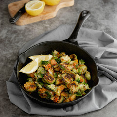 Roasted brussels sprouts in iron pan, with lemon slices. Dark background. Square image. Healthy food concept.