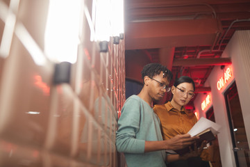 Waist up portrait of two ethnic business people discussing work documents while standing by wall outdoors in dim lighting, copy space
