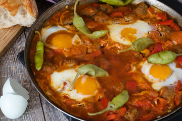 shakshuka in eiserner pfanne auf holztisch daneben ein schneidebrett mit brot pepperoni paprika tomaten und eierschalen