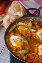shakshuka in eiserner pfanne auf holztisch daneben ein schneidebrett mit brot pepperoni paprika tomaten und eierschalen
