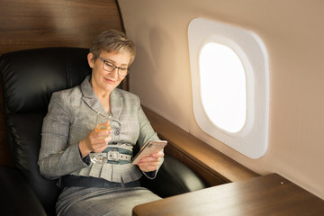 adult woman with a short haircut in a suit sits in the chair of a private plane with a phone and a...