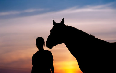 The girl near to a horse standing in front of a beautiful sunset. Silhouette of a woman and a horse's head.