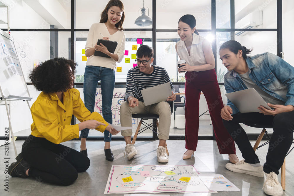 Wall mural Young creative asian group meeting and looking at project plan lay out on floor discuss or brainstorm business strategy with post note. Workshop for startup team in small office. Happy workplace.