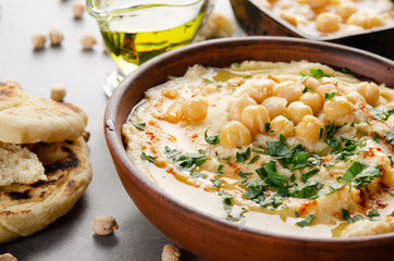 Hummus in clay dish topped with olive oil, chickpeas and green coriander leaves on stone table served with traditional Pita bread