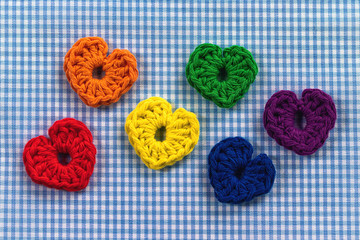 Six crocheted hearts. Rainbow colors. On light blue and white checkered fabric. 
