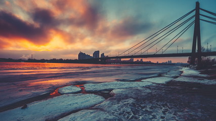 Orange grey evening clouds float over half frozen Dnipro river reflecting sunset by famous...