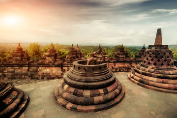 Amazing sunrise view of meditating Buddha statue and stone stupas against shining sun on background. Ancient Borobudur Buddhist temple. Great religious architecture. Magelang, Central Java, Indonesia