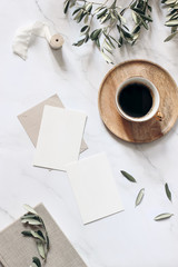 Summer wedding stationery mock-up scene. Blank greeting cards, wooden plate, book, ribbon, cup of coffee and olive branches. White table background with shadows. Vertical feminine flat lay, top view.