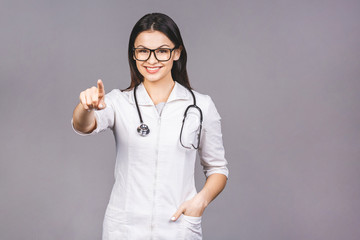 Portrait of cheerful young female doctor with stethoscope over neck looking at camera isolated on grey background. Pointing finger.