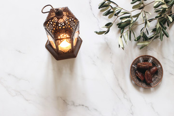Ramadan Kareem greeting card, invitation. Bronze plate with dates fruit, olive branches and glowing Moroccan lantern on white marble table. Iftar dinner. Eid ul Adha background. Flat lay, top view. © tabitazn