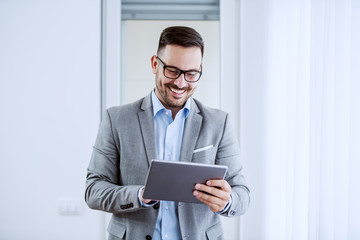 Attractive cheerful caucasian classy businessman in suit and with eyeglasses holding tablet and...