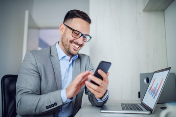 Handsome caucasian classy unshaven businessman in suit and with eyeglasses using smart phone while sitting in office. On table is laptop with chart on it.