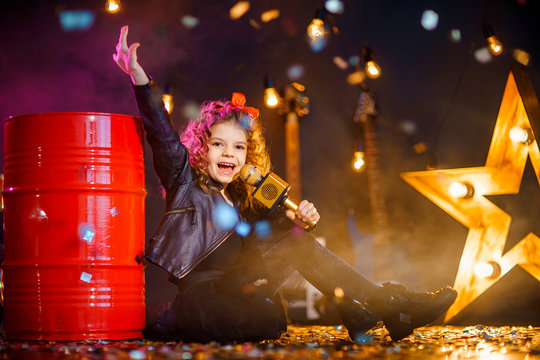 Beautiful girl with curly hair wearing leather jacket sing into a wireless microphone for karaoke while sitting on red tank in recording studio or stage.