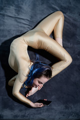 A female acrobat lies on a gray bedspread in a curved pose and uses a mobile. A woman contortionist...