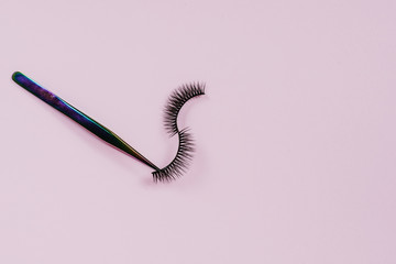 Rainbow tweezers hold false eyelashes on a pink pastel background. The concept of applying makeup. Copyspace, no one