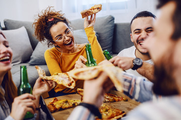 Group of friends sitting on the floor in living room, drinking beer and eating pizza. House party.