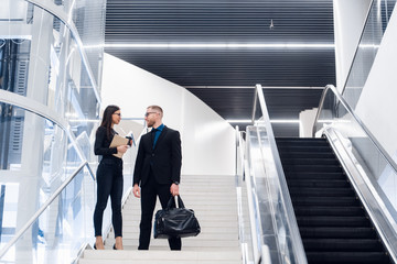 Business couple on a stairs talking and smiling. Man wearing suit and glasses with a bag talking with a woman holding tablet computer.