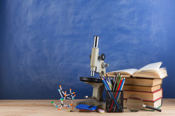 Education and sciences concept - books, molecule model and microscope on the desk in the auditorium, chalkboard background.