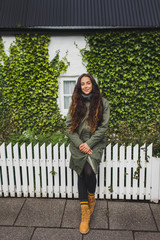 Curly brunette woman in khaki jacket and orange boots sitting on white fence of house with small white windows and wall, covered with green ivy plant. Trendy autumn look. © Oleg Breslavtsev