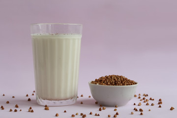 Glass beaker with milk on pink background. Next to the glass is a bowl of buckwheat, part of which is scattered on the table. Plant based milk concept.